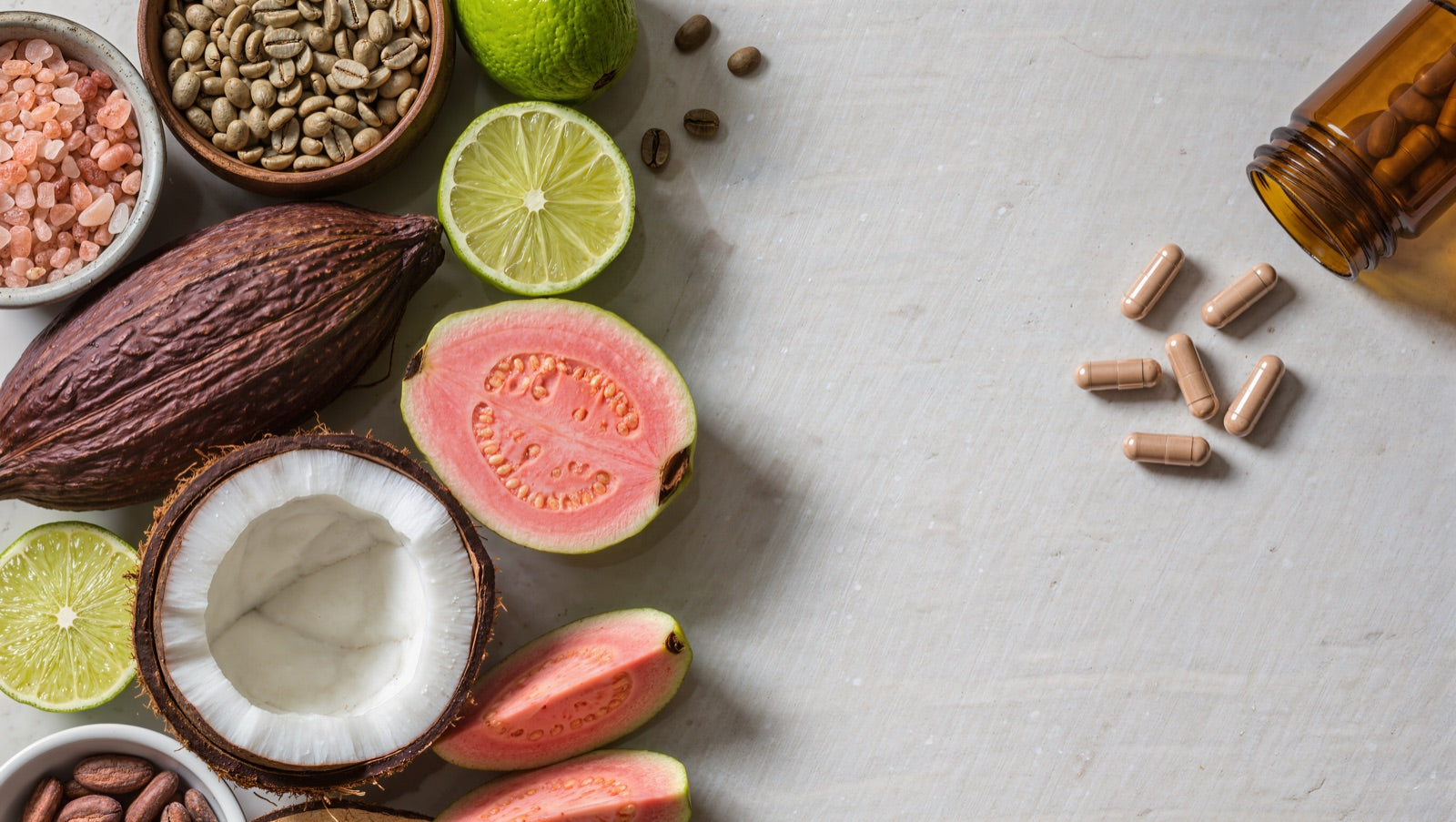 Fresh organic ingredients including cacao, coconut, guava, lime, green coffee beans and pink sea salt beside scattered beige supplement capsules and an amber pill bottle
