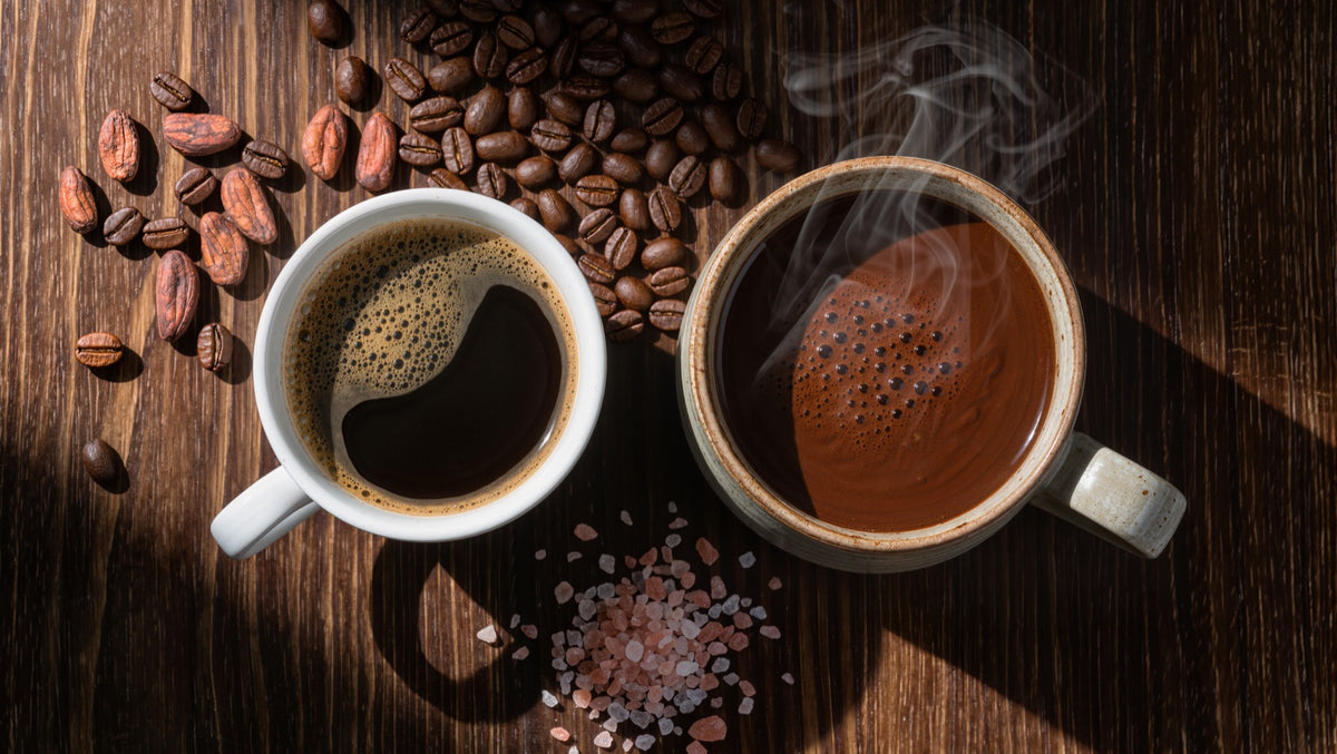A cup of black coffee beside a steaming mug of hot cacao with coffee beans, cacao nibs, and pink Himalayan salt on a wooden table